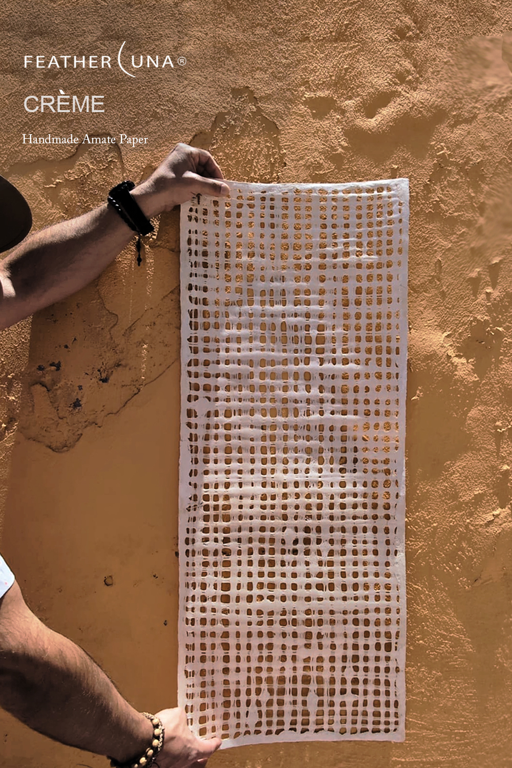 Hands holding a sheet of Crème handmade amate paper against a terracotta stucco wall, sunlight showing through its open grid