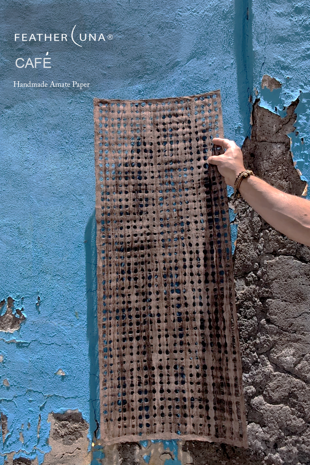 A hand holding a sheet of Café handmade amate paper against a weathered blue painted wall, sunlight showing through its open grid